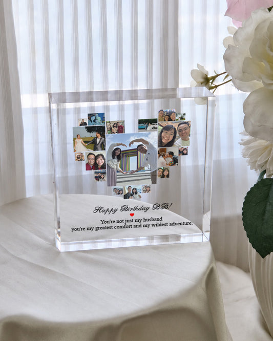 A clear acrylic photo plaque on a draped white fabric surface, featuring multiple small family photos arranged around a central portrait and a personalized birthday message. White flowers are visible at the right edge, and sheer curtains fill the background.